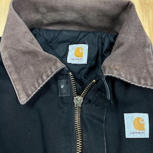 May include: Black work jacket with a brown corduroy collar and a brass zipper. The jacket features two white and orange Carhartt brand labels. The jacket is made of a durable material and is designed for work.