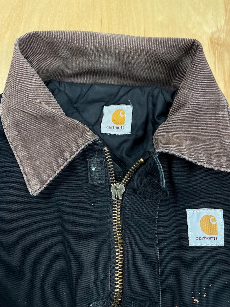 May include: Black work jacket with a brown corduroy collar and a brass zipper. The jacket features two white and orange Carhartt brand labels. The jacket is made of a durable material and is designed for work.