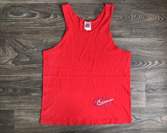 May include: Red sleeveless tank top with a white Nike swoosh logo on the front. The logo is surrounded by a blue circle.