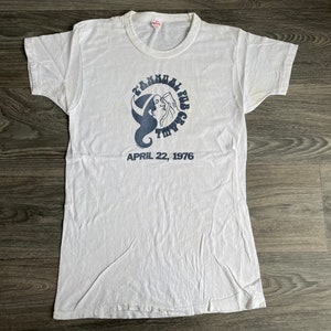 May include: White t-shirt with a blue graphic of a woman's face with the text "TAMMANY HALL FAB GEAR" and "APRIL 22, 1976" printed below.