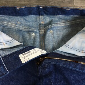 Puede incluir: Un par de pantalones vaqueros azules con cierre de cremallera y bot&oacute;n. Los vaqueros tienen una etiqueta Wrangler con el texto "Wrangler 13MWZ 32 x 34" en el interior de la cintura.