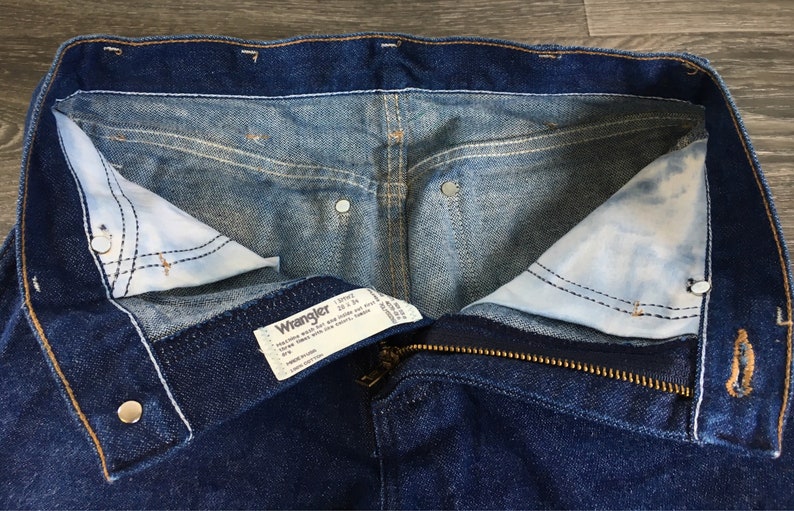 Puede incluir: Un par de pantalones vaqueros azules con cierre de cremallera y bot&oacute;n. Los vaqueros tienen una etiqueta Wrangler con el texto "Wrangler 13MWZ 32 x 34" en el interior de la cintura.