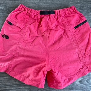 May include: Pink nylon shorts with black elastic waistband and black zippered pockets. The shorts have "The North Face" logo on the left leg.