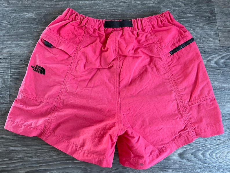 May include: Pink nylon shorts with black elastic waistband and black zippered pockets. The shorts have "The North Face" logo on the left leg.