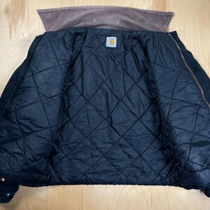 May include: A black quilted jacket with a brown corduroy collar. The jacket is unzipped, revealing a diamond-patterned interior lining. A small, square Carhartt label is visible inside the jacket.