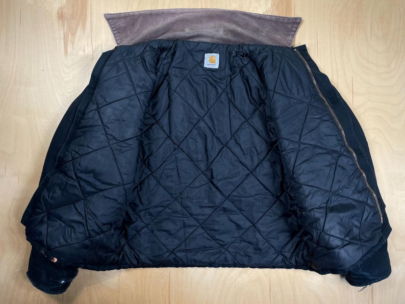 May include: A black quilted jacket with a brown corduroy collar. The jacket is unzipped, revealing a diamond-patterned interior lining. A small, square Carhartt label is visible inside the jacket.