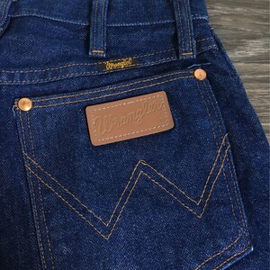 Puede incluir: Primer plano del bolsillo trasero de un par de pantalones vaqueros azules. El bolsillo tiene un parche de cuero marr&oacute;n con la palabra "Wrangler" bordada en blanco.