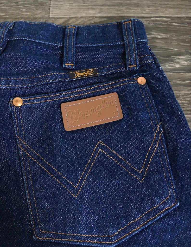 Puede incluir: Primer plano del bolsillo trasero de un par de pantalones vaqueros azules. El bolsillo tiene un parche de cuero marr&oacute;n con la palabra "Wrangler" bordada en blanco.