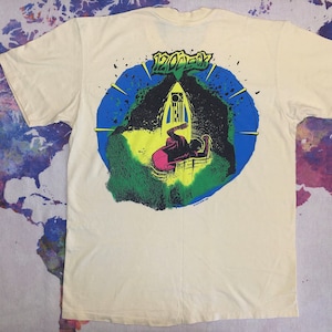 May include: A white t-shirt with a graphic print of a blue, green, and yellow circle with the text "1200ECK" in black. The graphic depicts a person in a pink shirt and black pants standing in front of a building with a yellow door.