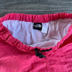 May include: Pink drawstring pants with a black drawstring and a white label that says "The North Face" in black text.
