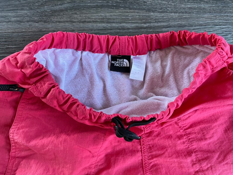 May include: Pink drawstring pants with a black drawstring and a white label that says "The North Face" in black text.