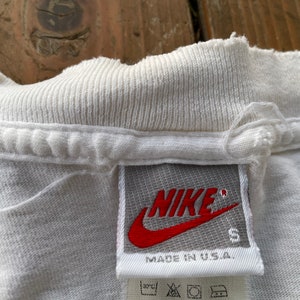 K&ouml;nnte beinhalten: Wei&szlig;es T-Shirt mit einem grauen Nike-Etikett, auf dem "NIKE" in Rot mit einem roten Swoosh-Logo und "MADE IN U.S.A." in Schwarz steht. Das Etikett tr&auml;gt auch die Gr&ouml;&szlig;e "S".