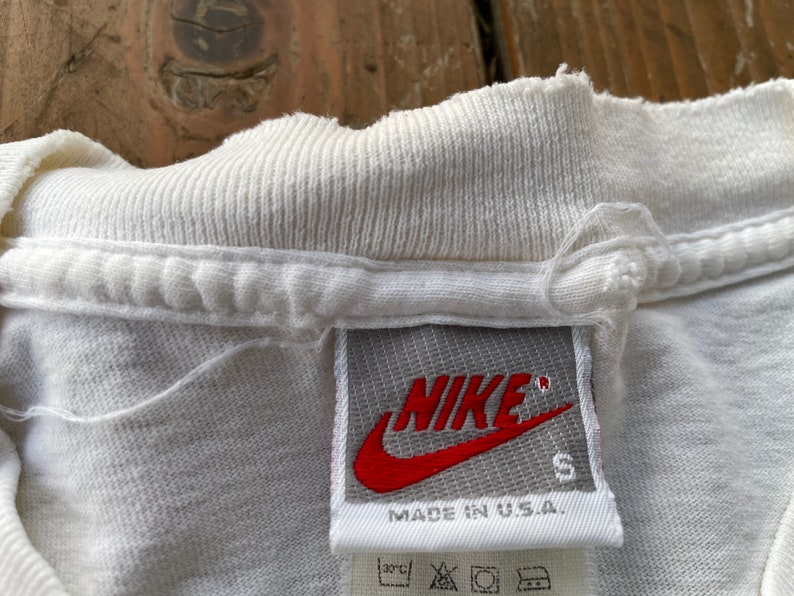 K&ouml;nnte beinhalten: Wei&szlig;es T-Shirt mit einem grauen Nike-Etikett, auf dem "NIKE" in Rot mit einem roten Swoosh-Logo und "MADE IN U.S.A." in Schwarz steht. Das Etikett tr&auml;gt auch die Gr&ouml;&szlig;e "S".