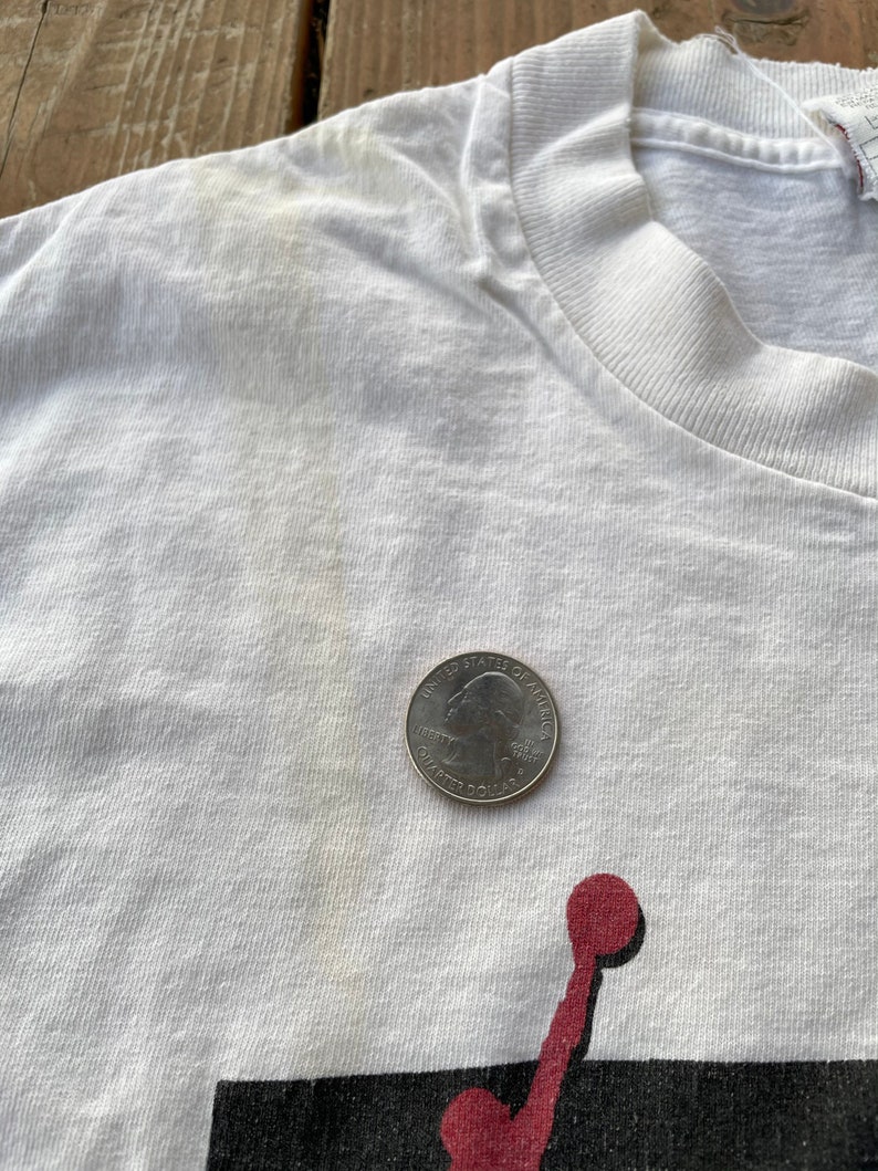 K&ouml;nnte beinhalten: Wei&szlig;es T-Shirt mit einem roten und schwarzen Grafikdruck eines Basketballspielers. Das Shirt hat einen verblassten Look und eine 1-Euro-M&uuml;nze liegt zum Gr&ouml;&szlig;envergleich auf dem Shirt.