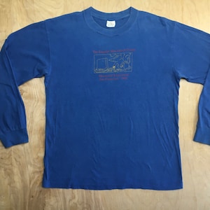 May include: A blue long-sleeve t-shirt with a yellow graphic of a cartoon scene and the text "The Chronicle of Higher Education. May we suggest a reading list? San Francisco, CA 1984."