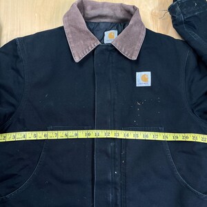 May include: Black Carhartt jacket with a brown corduroy collar. The jacket has a button-up front, two front pockets, and a Carhartt logo patch. A yellow measuring tape is laid across the jacket, measuring approximately 25 inches across.