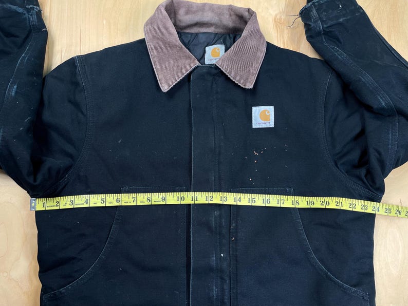 May include: Black Carhartt jacket with a brown corduroy collar. The jacket has a button-up front, two front pockets, and a Carhartt logo patch. A yellow measuring tape is laid across the jacket, measuring approximately 25 inches across.
