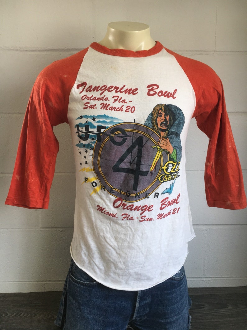 May include: A white and orange baseball t-shirt with a graphic of a clock and a person holding a banana. The text on the shirt reads "Tangerine Bowl", "Orlando, Fla. - Sat. March 20", "FOREIGNER", "Orange Bowl", "Miami, Fla. - Sun. March 21".