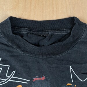 May include: Black t-shirt featuring a blue and orange cartoon character wearing a blue hat and holding a sword. The character has red hair and a white outline. The text "M" is visible in the top right corner of the image.