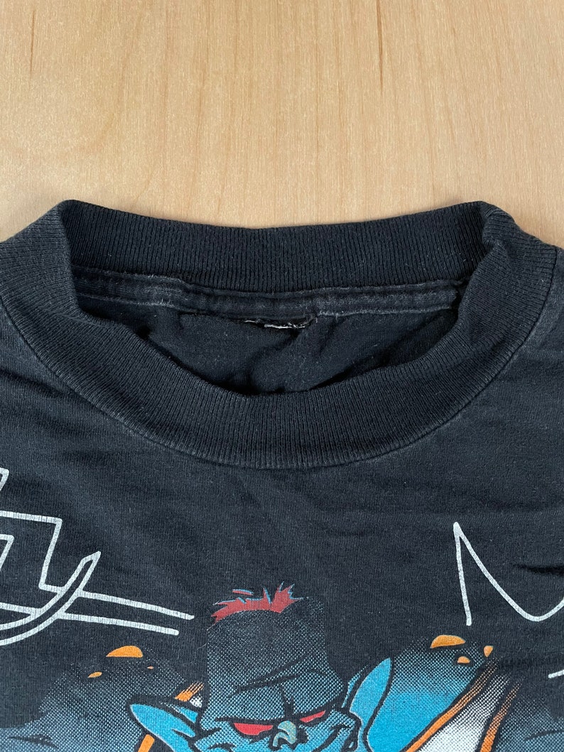 May include: Black t-shirt featuring a blue and orange cartoon character wearing a blue hat and holding a sword. The character has red hair and a white outline. The text "M" is visible in the top right corner of the image.