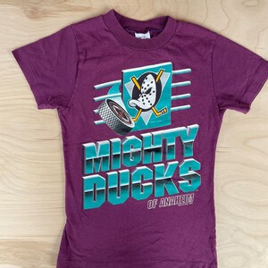 Puede incluir: Una camiseta morada con un logotipo de los Mighty Ducks de Anaheim en turquesa y blanco. El logotipo presenta un pato de dibujos animados con una máscara de hockey y un palo de hockey.