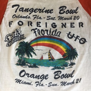 May include: A white t-shirt with a red and orange baseball sleeve design. The shirt features a rainbow, palm trees, and sailboats. The text on the shirt reads "Tangerine Bowl Orlando, Fla. - Sat. March 20 FOREIGNER Florida UFO Orange Bowl Miami, Fla. - Sun. March 21".