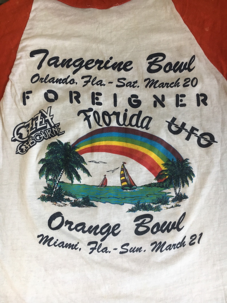 May include: A white t-shirt with a red and orange baseball sleeve design. The shirt features a rainbow, palm trees, and sailboats. The text on the shirt reads "Tangerine Bowl Orlando, Fla. - Sat. March 20 FOREIGNER Florida UFO Orange Bowl Miami, Fla. - Sun. March 21".