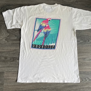 May include: White t-shirt with a graphic of two red, blue, and yellow parrots on a green and blue background. The graphic is framed in a black rectangle with the word "PARADISE" printed in white below the parrots.