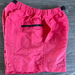 May include: Pink nylon shorts with a black elastic waistband and a zippered pocket on the right side.