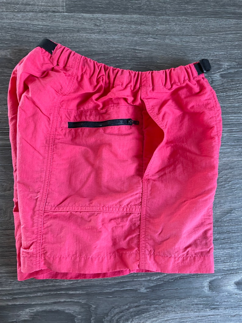 May include: Pink nylon shorts with a black elastic waistband and a zippered pocket on the right side.