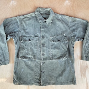 Può includere: Una camicia da lavoro verde con bottoni e due grandi tasche. La camicia è realizzata in tessuto di cotone resistente e ha un aspetto sbiadito e vintage.