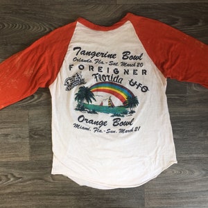 May include: A white and orange baseball style t-shirt with a graphic of a rainbow over a beach scene. The text on the shirt reads "Tangerine Bowl Orlando, Fla. - Sat. March 20 FOREIGNER Florida UFO Orange Bowl Miami, Fla. - Sun. March 21"