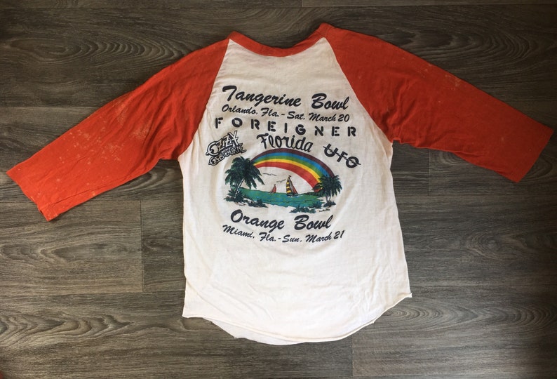 May include: A white and orange baseball style t-shirt with a graphic of a rainbow over a beach scene. The text on the shirt reads "Tangerine Bowl Orlando, Fla. - Sat. March 20 FOREIGNER Florida UFO Orange Bowl Miami, Fla. - Sun. March 21"