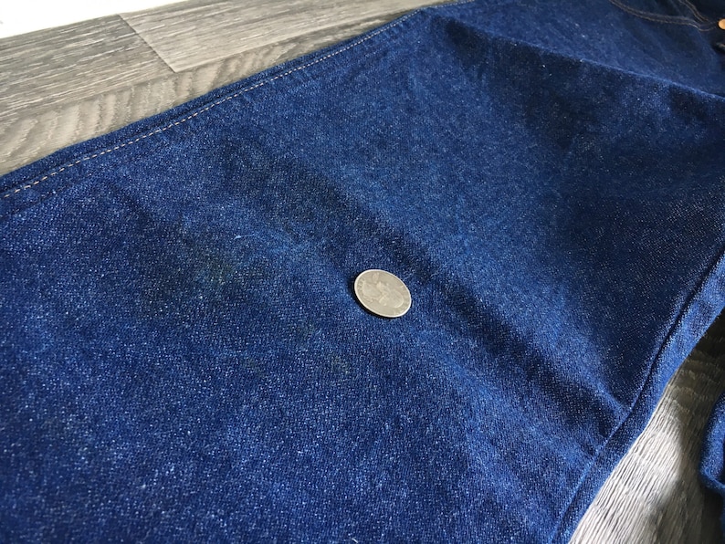 Puede incluir: Primer plano de tela vaquera azul oscuro con una textura sutil. Una moneda de plata se coloca sobre la tela para la escala.