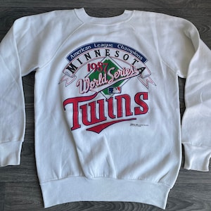 Peut inclure: Sweat-shirt blanc avec un imprimé graphique rouge, bleu et vert qui dit "American League Champions Minnesota 1987 World Series Twins".