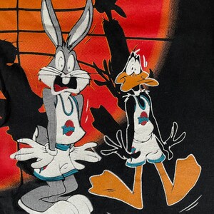 May include: Bugs Bunny and Daffy Duck are wearing basketball uniforms and look scared. They are standing on a black background with an orange and red sunset in the background.