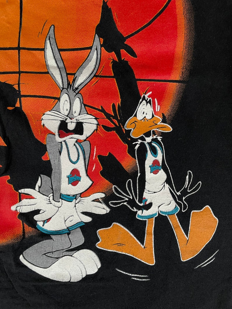 May include: Bugs Bunny and Daffy Duck are wearing basketball uniforms and look scared. They are standing on a black background with an orange and red sunset in the background.