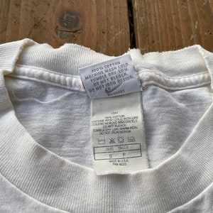 K&ouml;nnte beinhalten: Wei&szlig;es Baumwoll-T-Shirt mit einem Pflegeetikett, auf dem steht "100% Baumwolle, Maschinenw&auml;sche kalt, Trocknergeeignet bei niedriger Temperatur, Nicht bleichen, Nicht auf das Design b&uuml;geln". Das Etikett enth&auml;lt auch die Gr&ouml;&szlig;e "S" und den Text "Hergestellt in den USA, RN# 56323".