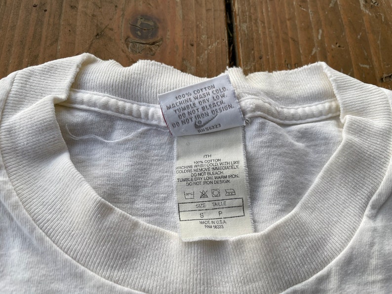K&ouml;nnte beinhalten: Wei&szlig;es Baumwoll-T-Shirt mit einem Pflegeetikett, auf dem steht "100% Baumwolle, Maschinenw&auml;sche kalt, Trocknergeeignet bei niedriger Temperatur, Nicht bleichen, Nicht auf das Design b&uuml;geln". Das Etikett enth&auml;lt auch die Gr&ouml;&szlig;e "S" und den Text "Hergestellt in den USA, RN# 56323".