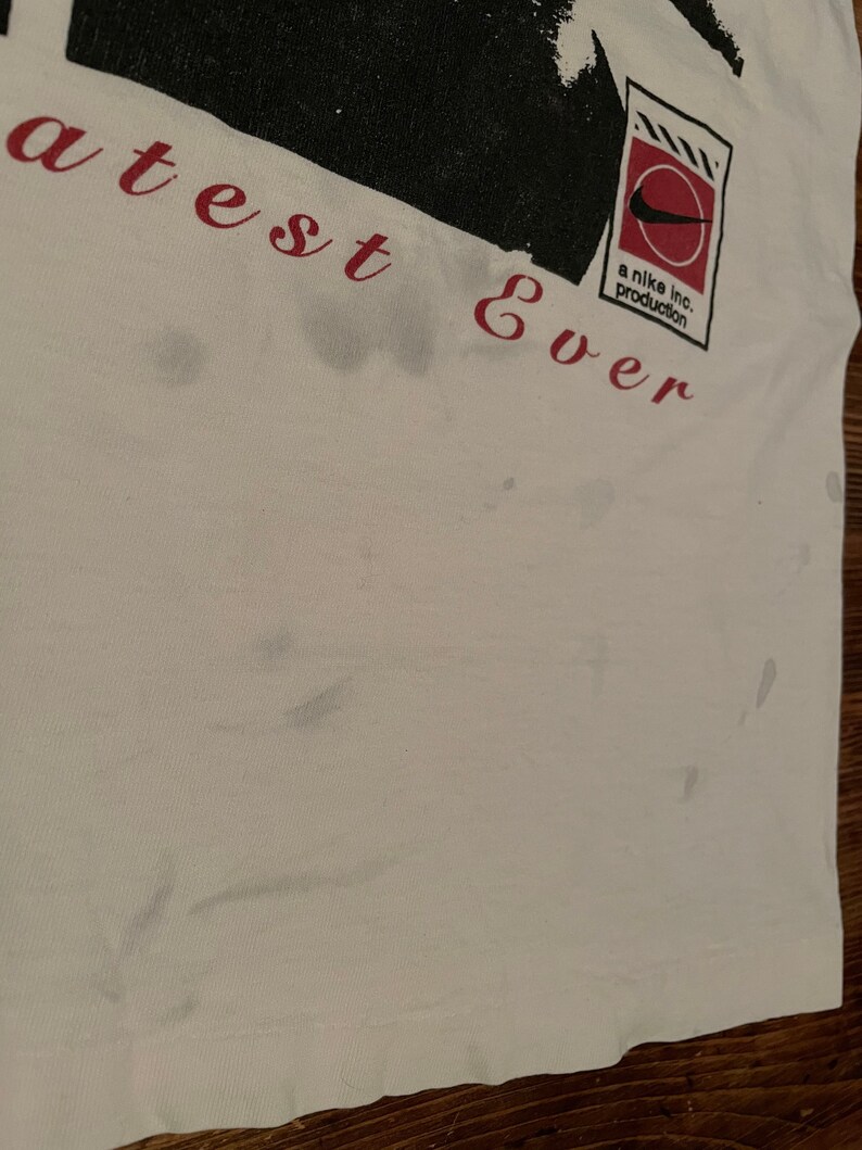 K&ouml;nnte beinhalten: Wei&szlig;es T-Shirt mit einer schwarz-wei&szlig;en Grafik einer Person mit dem Text "greatest ever" und einem rot-wei&szlig;en Nike-Logo mit dem Text "a nike inc. production".