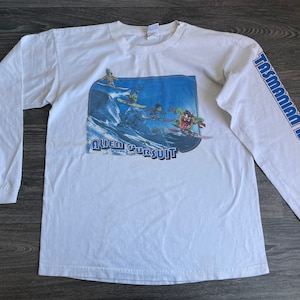 Op de afbeelding: Witte longsleeve t-shirt met een cartoon afbeelding van de Tasmaanse Duivel die surft met andere personages. De tekst "Alien Pursuit" is onder de afbeelding gedrukt. De woorden "Tasmanian Devil" staan verticaal op de rechtermouw gedrukt.