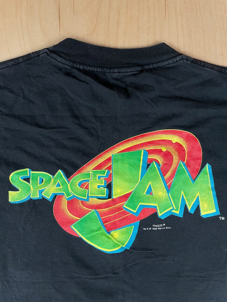 May include: Black t-shirt with a green and red Space Jam logo featuring the words "Space Jam" and the Warner Bros. "Freeze" logo.