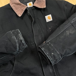 May include: Black work jacket with a brown corduroy collar. The jacket has a zipper closure and a Carhartt logo patch. The sleeves show signs of wear and paint marks. A durable and functional outerwear piece.