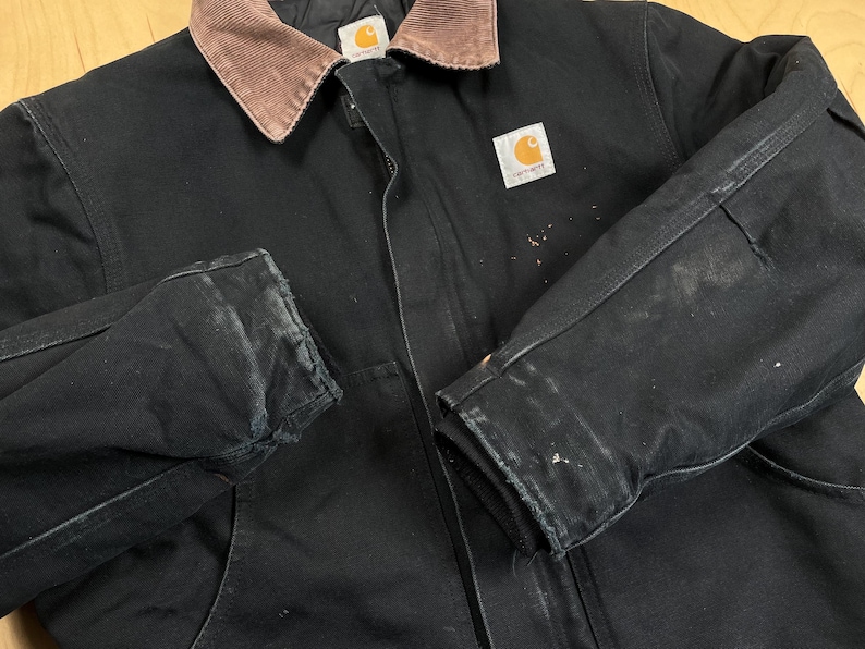 May include: Black work jacket with a brown corduroy collar. The jacket has a zipper closure and a Carhartt logo patch. The sleeves show signs of wear and paint marks. A durable and functional outerwear piece.