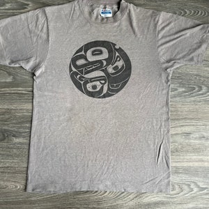 May include: A grey t-shirt with a black and white graphic print of a stylised design. The design is a circle with a pattern of stylised figures.