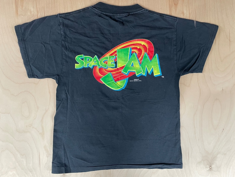May include: Black t-shirt with a colourful Space Jam logo on the back. The logo features the words "Space Jam" in a stylised font with a red, yellow and green basketball hoop graphic.