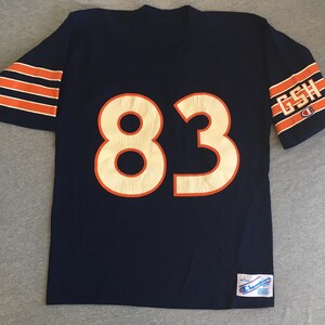 Pode incluir: Uma camisa de futebol azul marinho com listras laranja e brancas nas mangas. O número "83" está na frente da camisa em branco com contorno laranja. A palavra "GSH" está na manga direita em branco com contorno laranja. A camisa tem um logotipo da Champion na manga direita e uma etiqueta da Champion na parte inferior direita.