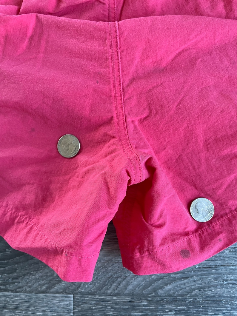 May include: Close-up of a pair of pink shorts with two US quarters placed on the fabric. The shorts have a seam down the center back.