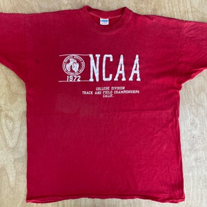 Puede incluir: Camiseta roja con estampado blanco. El estampado presenta un logotipo con el texto "NCAA 1972 College Division Track and Field Championships Calif."