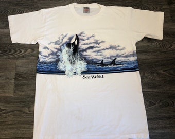 May include: White t-shirt with a graphic of an orca jumping out of the water with a blue and white background. The text "Sea World" is printed below the graphic.